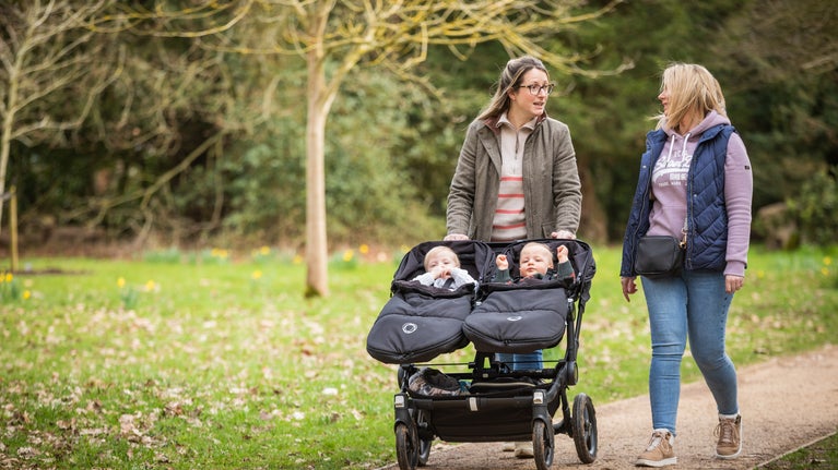 Two women walking down a path. One is pushing a stroller with two babies in.
